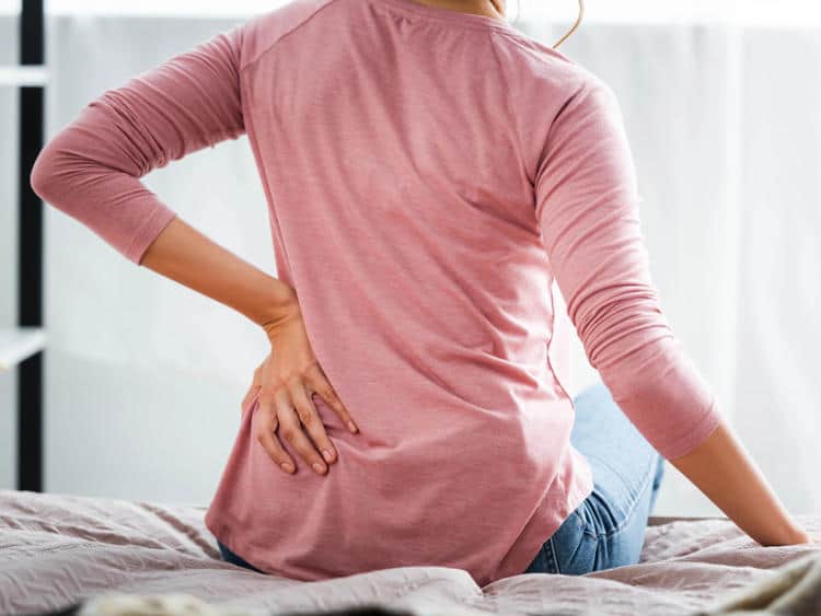 Woman experiencing back pain while sitting on a mattress.