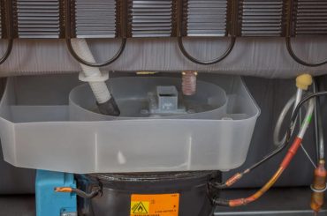 Rear view of a refrigerator showing the compressor and the area where refrigerant lines and service ports are located.