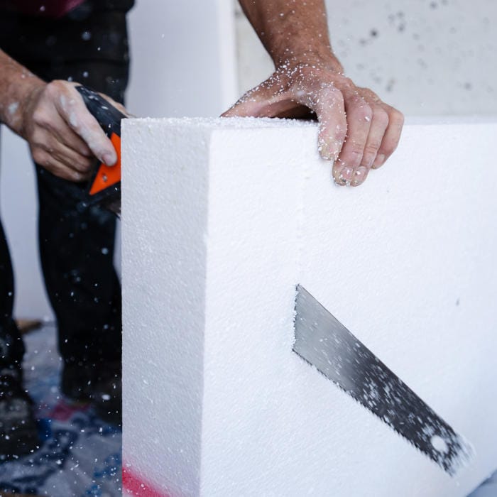 Worker cutting EPS insulation board with a handsaw during floor or wall insulation installation.