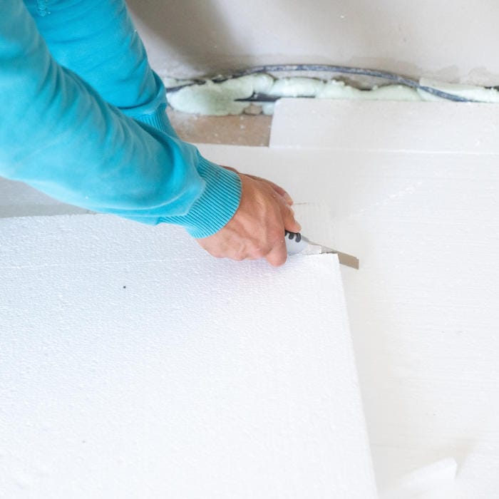 A person cutting rigid polystyrene foam insulation using a utility knife to achieve a clean, precise fit during floor installation.